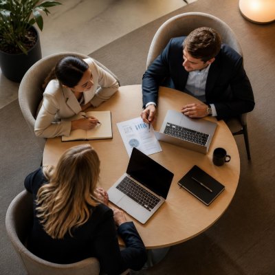 Three colleagues seen from above, working around a small round table with two laptops, a printed chart between them and one of them taking notes.